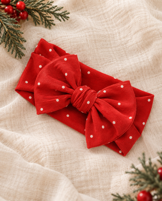 Shae & Palmer Ruby Dot bamboo baby bow headband featuring a festive red and white polka dot print, styled on soft cream linen with evergreen branches for a cozy Christmas holiday flat lay.