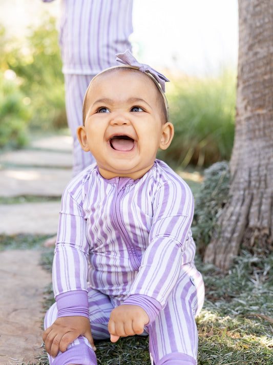 Smiling baby in lavender striped bamboo pajamas outdoors, sitting on a garden path with a matching family member in the background