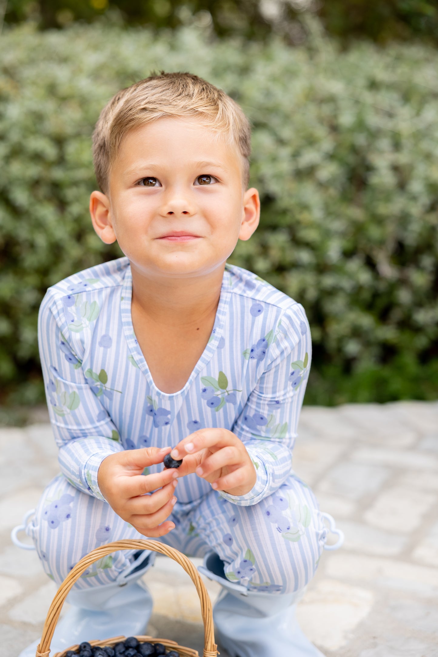 Toddler boy in Blueberry Lane blue striped bamboo pajamas holding a fresh blueberry beside a berry basket, soft and breathable sleepwear for sensitive skin
