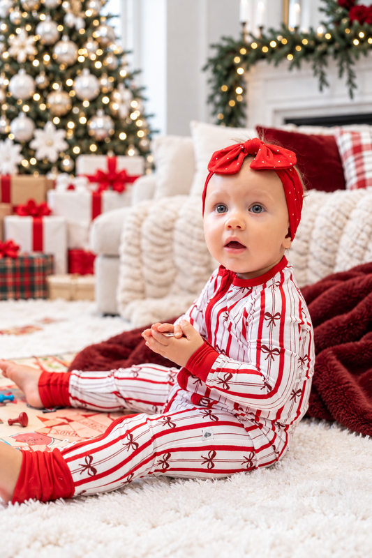 Baby wearing red and white Christmas pajamas with bow print and red polka dot headband, surrounded by Christmas decorations, tree and presents