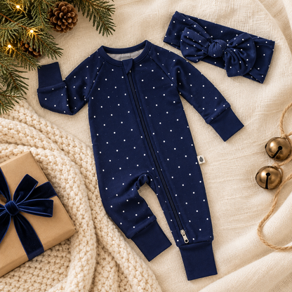 Shae & Palmer Midnight Dot navy blue bamboo baby romper with white polka dots, double zipper, solid navy cuffs, and matching baby bow headband. Holiday flat lay styled with cream blankets, greenery, and wrapped gifts for Christmas and Hanukkah.