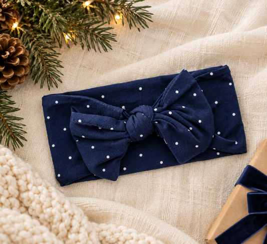 Shae & Palmer Midnight Dot navy blue bamboo baby bow headband featuring white polka dots, styled on soft cream linen with evergreen branches, pinecones, and holiday decorations. A festive baby accessory for Christmas and Hanukkah outfits.