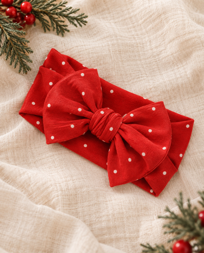 Shae & Palmer Ruby Dot bamboo baby bow headband featuring a festive red and white polka dot print, styled on soft cream linen with evergreen branches for a cozy Christmas holiday flat lay.