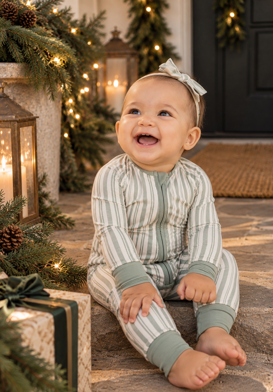 Smiling baby girl wearing Shae & Palmer's Studio Stripe Green bamboo zip-up romper with matching bow, sitting on stone steps surrounded by festive greenery, twinkling lights, and holiday gifts. Cozy, soft bamboo baby pajamas perfect for Christmas and Hanukkah.