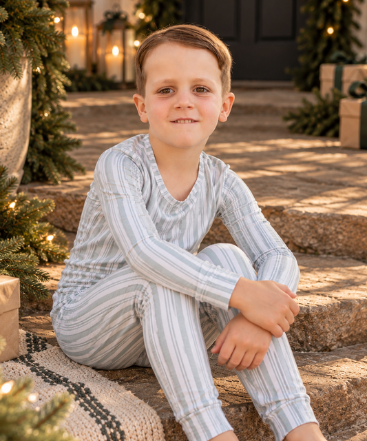 Young boy wearing Shae & Palmer Studio Stripe Green bamboo big kids two-piece pajamas, sitting on stone steps surrounded by festive evergreen garlands, glowing lanterns, and holiday gifts. Soft, snug-fitting striped pajamas for cozy Christmas and Hanukkah mornings.