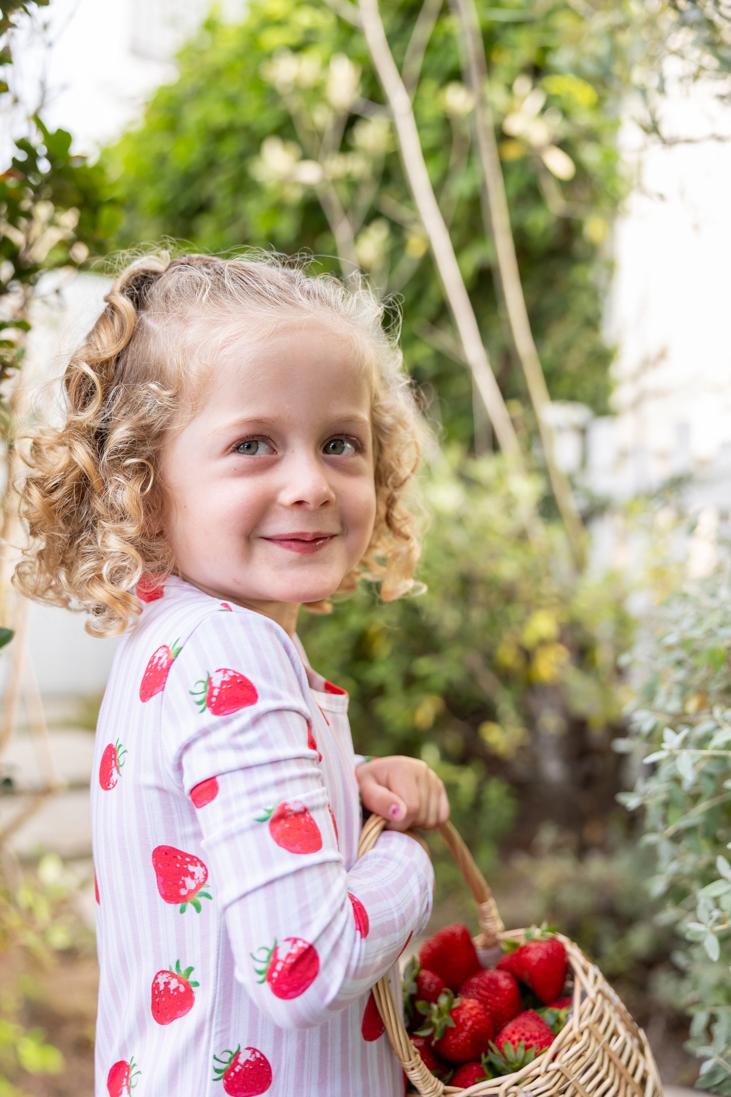 Toddler girl in pink strawberry-print bamboo pajama set holding a basket of fresh strawberries in a garden