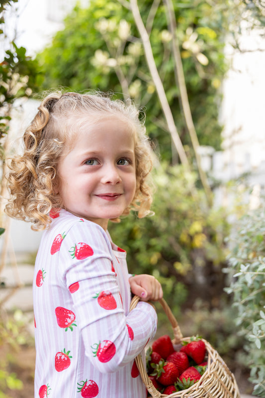 Toddler girl in pink strawberry-print bamboo pajama set holding a basket of fresh strawberries in a garden