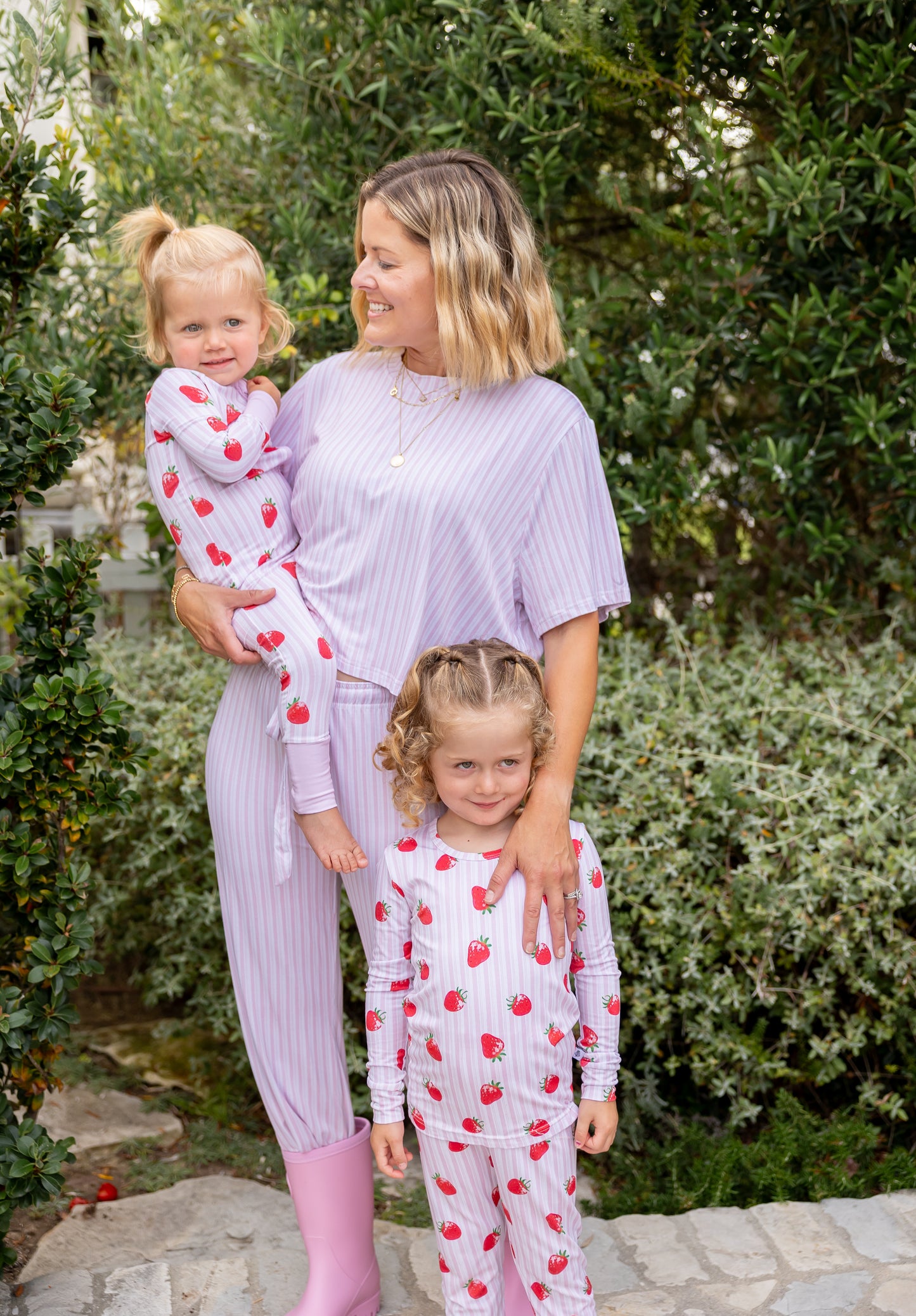 Mom in Strawberry stripe pink striped bamboo pajamas holding a toddler, with a big kid daughter nearby in matching strawberry print, part of Shae & Palmer's Market Fresh mommy-and-me collection
