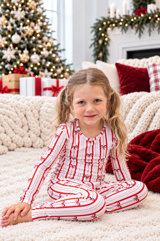 Toddler girl wearing red and white Ribbon Stripe bamboo pajamas with bow print in a cozy Christmas setting with Christmas tree, gifts, and holiday decor.